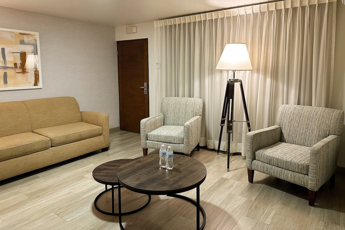 A hotel room with a sofa, two armchairs, a small round table with water bottles, a floor lamp, and a painting on the wall.