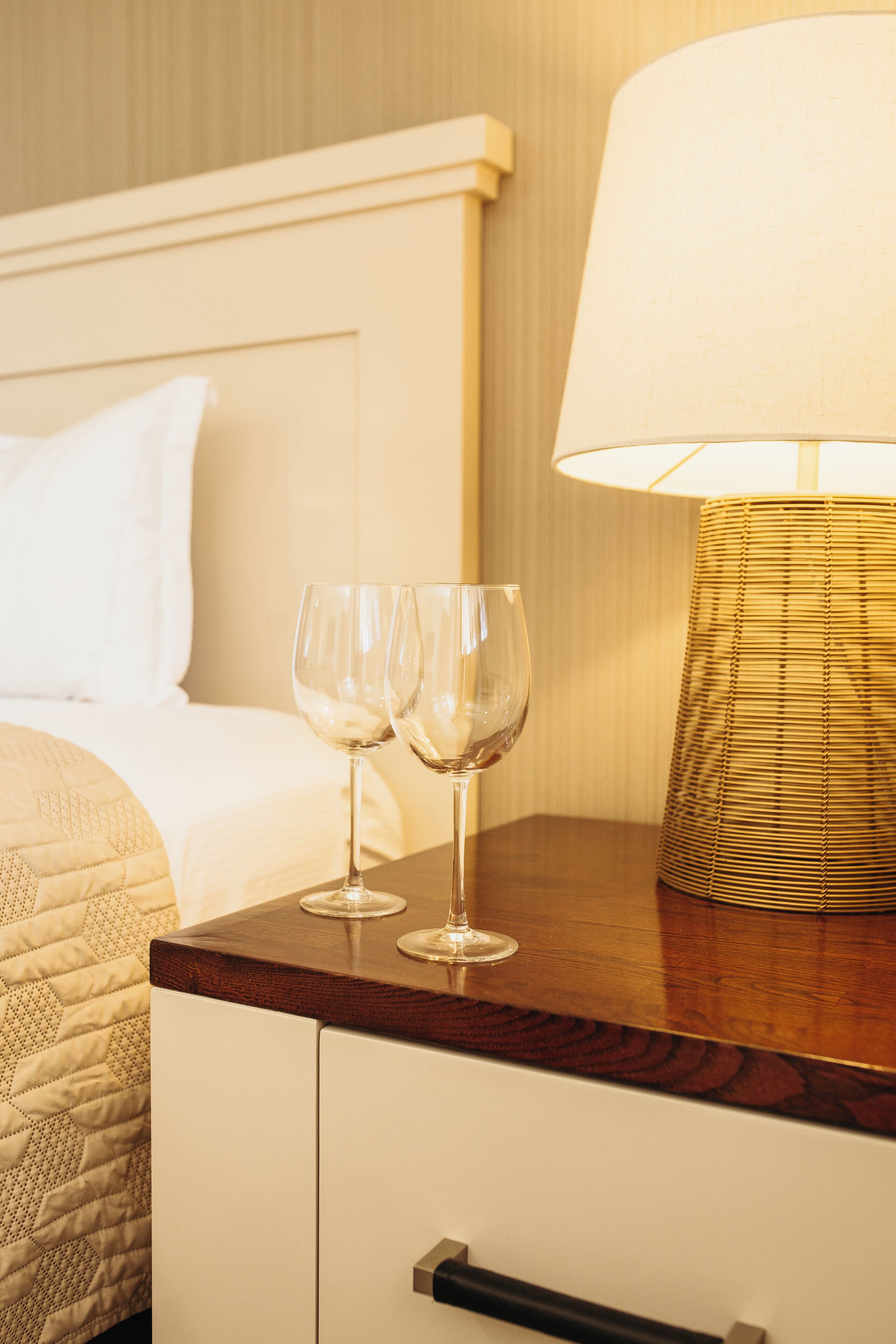 Un lit bien fait, avec une tête de lit blanche, une table de chevet en bois sur laquelle reposent deux verres à vin, et une lampe dotée d’un abat-jour en osier.