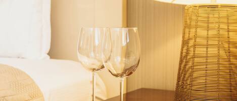 Un lit bien fait, avec une tête de lit blanche, une table de chevet en bois sur laquelle reposent deux verres à vin, et une lampe dotée d’un abat-jour en osier.