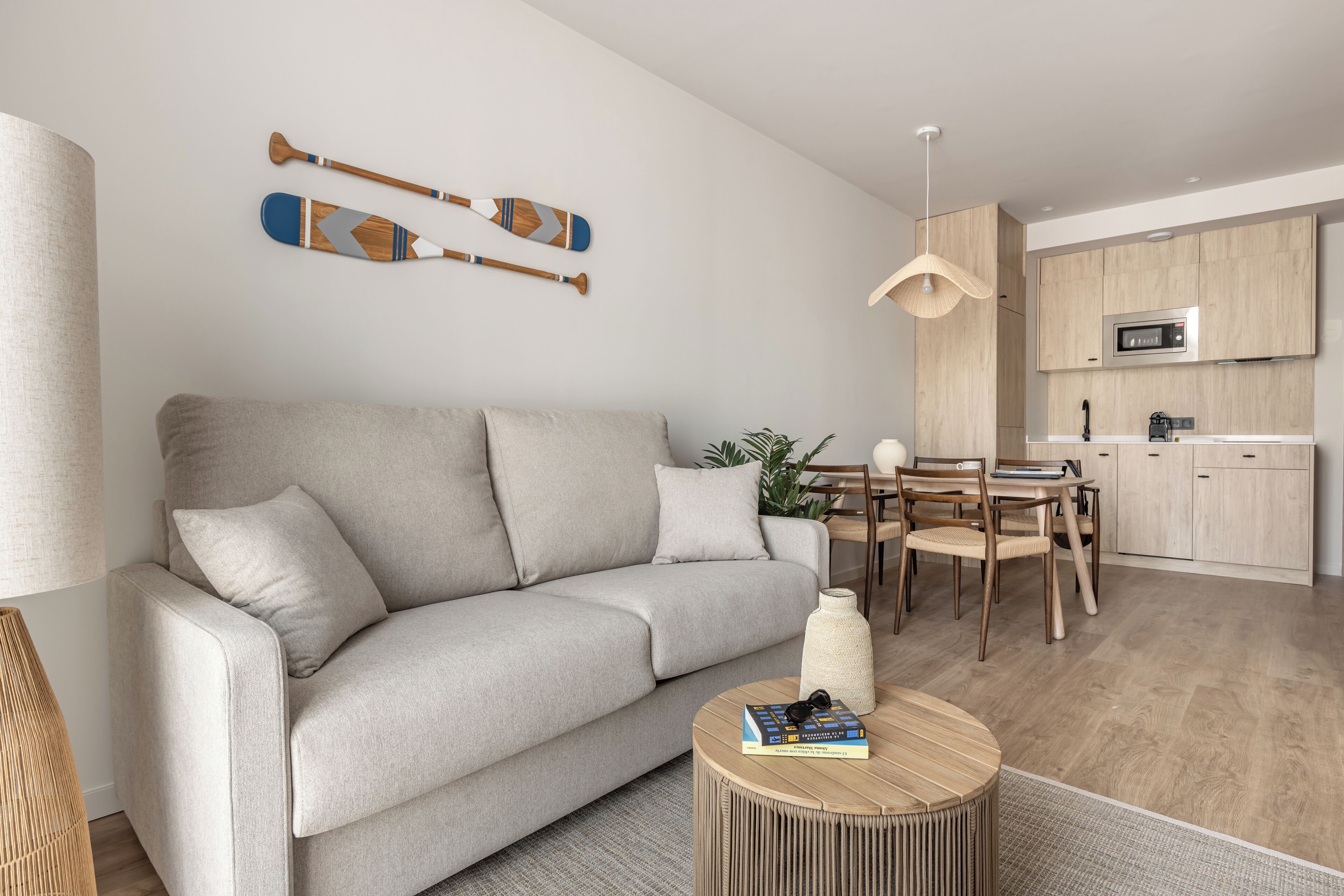 A modern living room with a grey sofa, wooden dining table, and a kitchen area with wooden cabinets and a microwave.