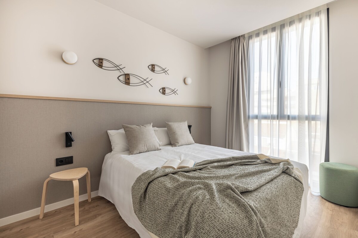 A modern bedroom with a bed, a side table, a green ottoman, and abstract wall art.