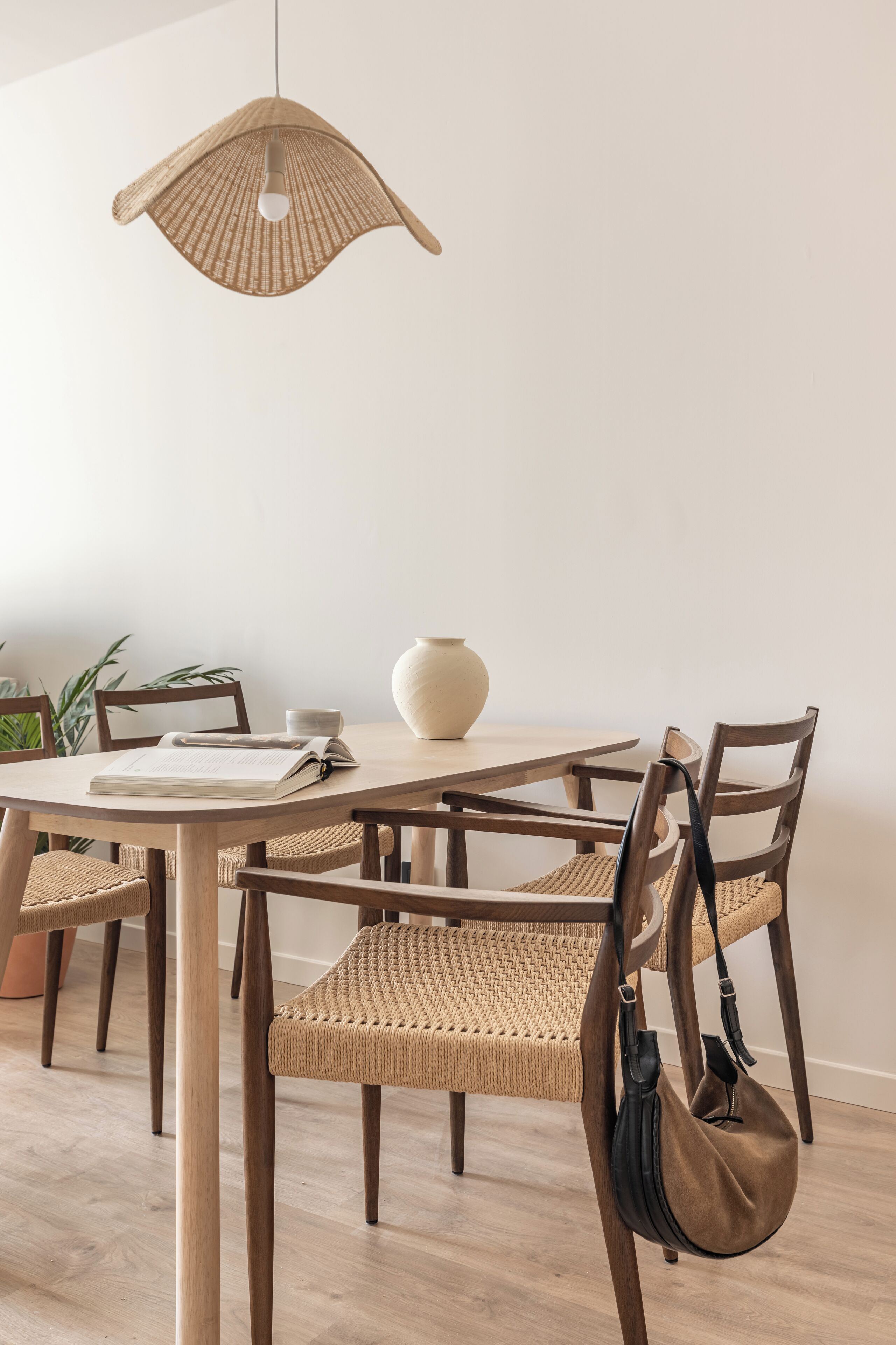 A dining area with a round table, wooden chairs, a woven chair, a hanging light fixture, and a vase on the table.
