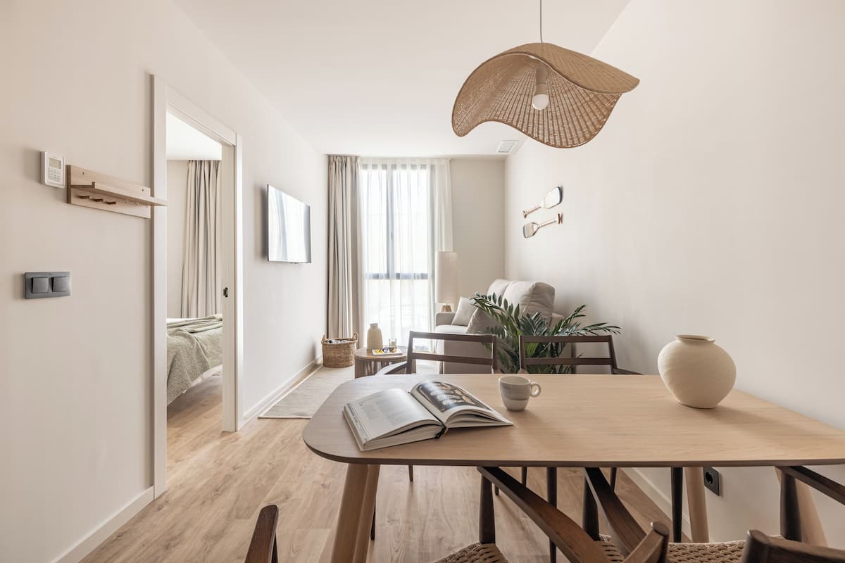 A modern, well-lit room with a wooden dining table, chairs, a hanging light fixture, and a view into another room with a bed and curtains.
