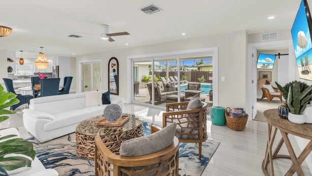 A spacious living room with a white sofa, wicker chairs, and a coffee table. A large window offers a view of a pool and outdoor furniture. - Stunning 4 Bed I Heated Pool/Hot Tub (Marco Island)