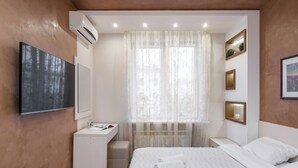 Een moderne slaapkamer met een bed, een bureau, een kaptafel en een groot raam met dunne gordijnen.