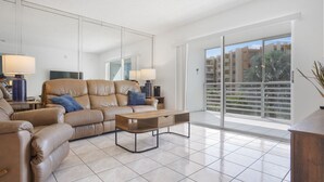A spacious living room with a large sofa, a coffee table, and a TV. There is a balcony with a view of buildings and trees. - Charming 2-bedroom condo nearby the beach with WiFi, AC in Sunny Isles Beach (Sunny Isles Beach)