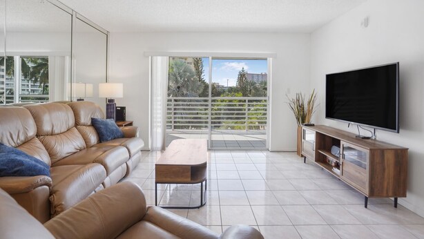 A modern living room with a leather sofa, a coffee table, a TV on a wooden stand, and a balcony with a view of palm trees and buildings. - Charming 2-bedroom condo nearby the beach with WiFi, AC in Sunny Isles Beach (Sunny Isles Beach)