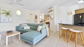 Ein modernes Wohnzimmer mit einem Ecksofa, einem kleinen Tisch und einer Küchenzeile mit Kühlschrank und Barhocker.