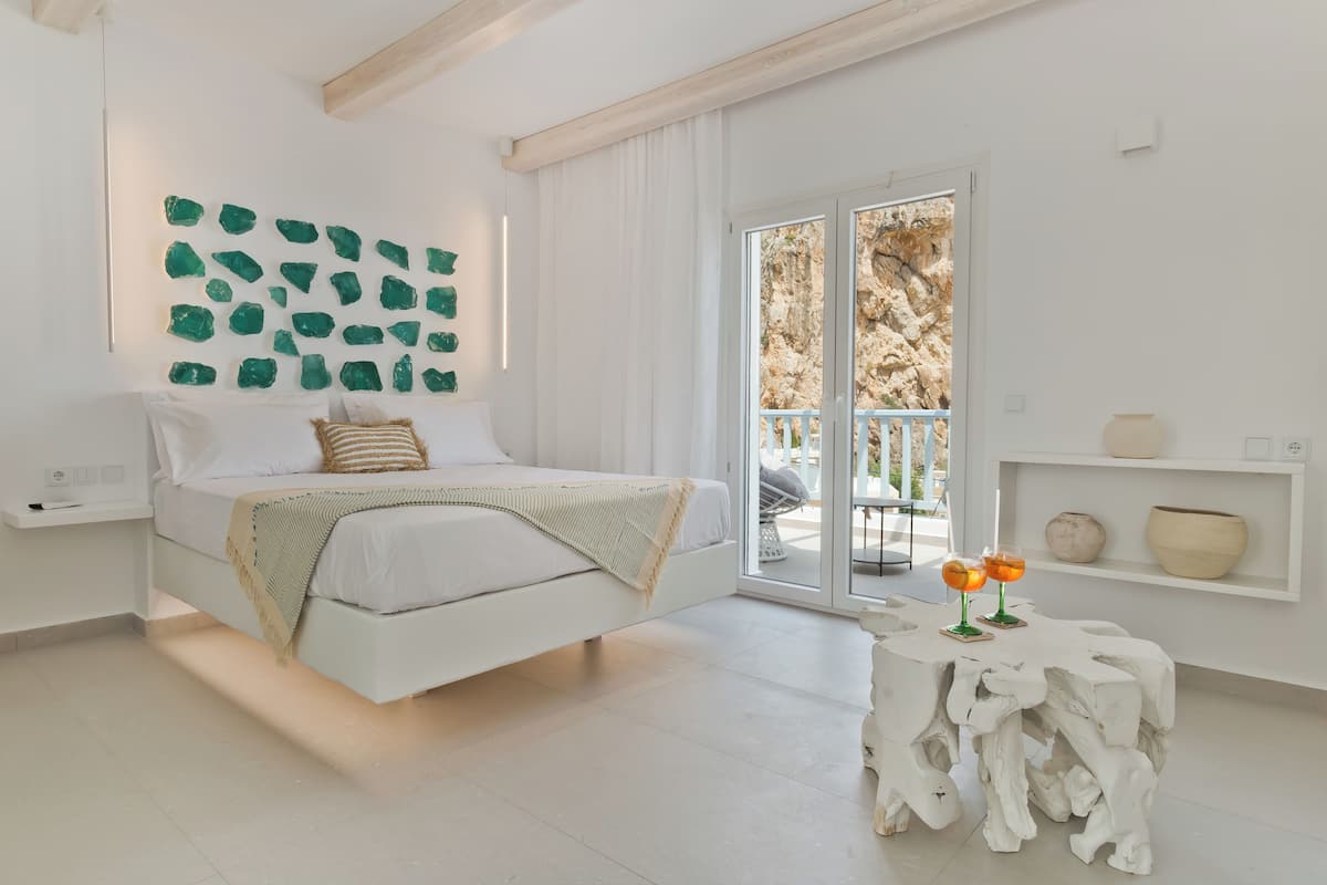 A modern bedroom with a bed, a wall-mounted green mineral art piece, a white bedside table with two glasses, and a view of the outdoors through a sliding glass door.