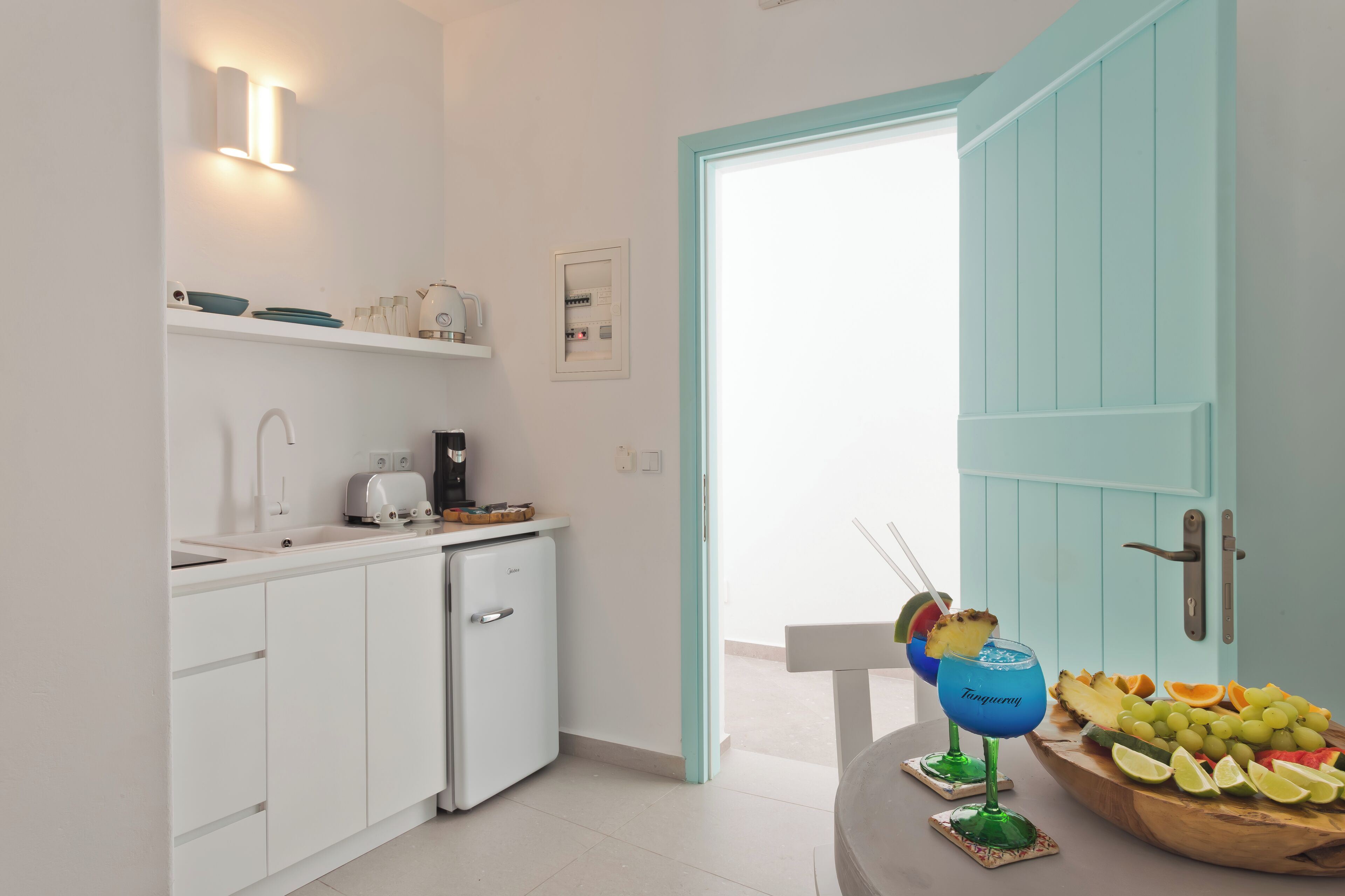 A modern kitchen with white cabinets, a small fridge, and a coffee machine. There is a wooden table with a fruit bowl and two drinks.
