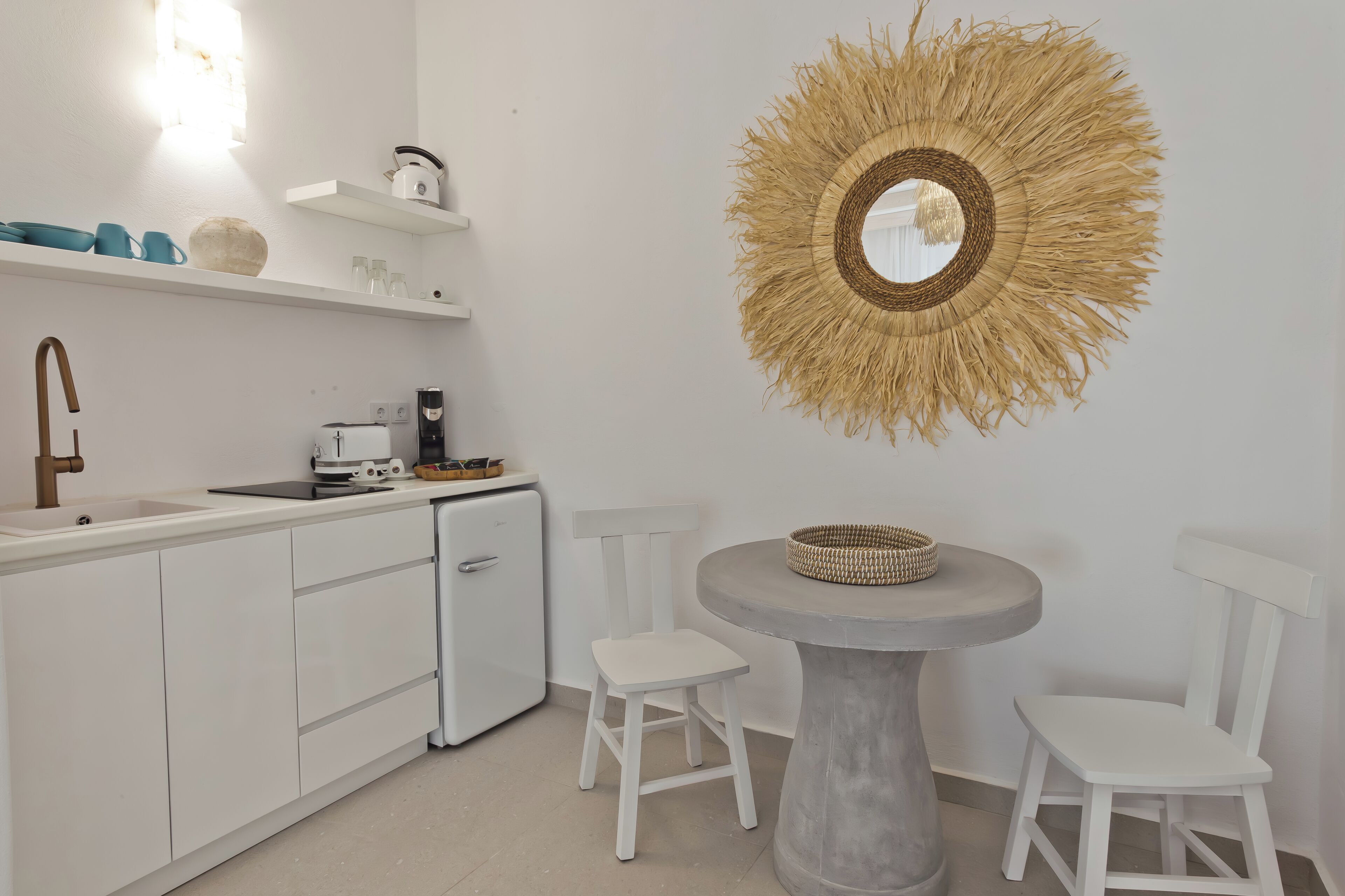 A modern kitchen with white cabinetry, a built-in dishwasher, and a small refrigerator. There is a round table with two white chairs and a woven wall hanging above it.