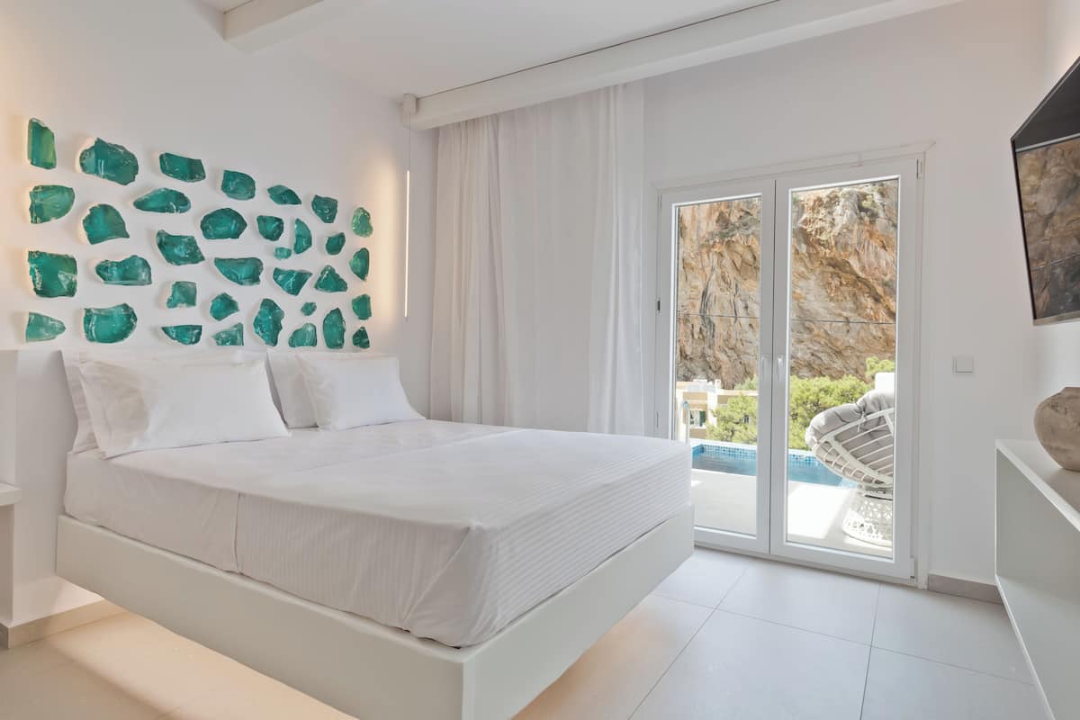A modern bedroom with a white bed, a large window with a view of a pool and greenery, and a wall decorated with green stones.