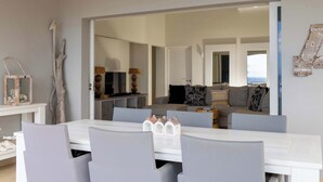 Ein Essbereich mit einem weißen Tisch und grauen Stühlen, ein Spiegel, der ein Wohnzimmer mit einer Couch und Kissen reflektiert, und ein dekoratives Element an der Wand.