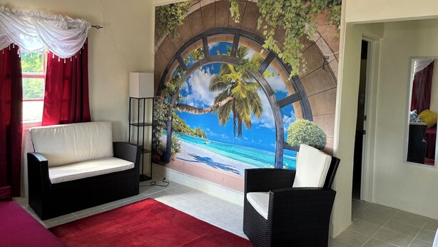 Una habitación con un gran mural de una playa tropical, un sofá blanco, una silla de mimbre negro y una alfombra roja.