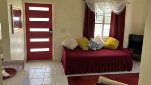 Una habitación con puerta roja, cama con colcha roja y varias almohadas, televisor y ventana con cortinas rojas.