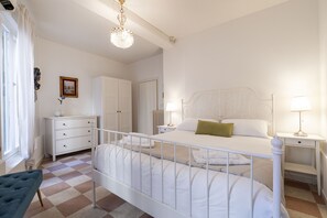 Een slaapkamer met een bed, nachtkastjes, een dressoir, een kroonluchter en een raam met gordijnen.