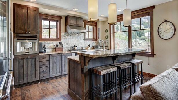 A kitchen with wooden cabinets, a large island with bar stools, a stainless steel range hood, and a view of a landscape through the windows. - Luxury 4 Bedroom Condo on Peak 8 4 Condo (Breckenridge)
