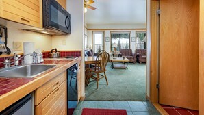 A compact kitchen with wooden cabinets, a microwave, and a sink. A dining area with a table and chairs is visible in the background. - 2797 Slopeside Condo (Keystone)