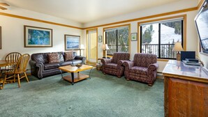 A living room with a brown leather sofa, patterned armchairs, a wooden coffee table, and a desk with a computer. Large windows offer a view of a snowy landscape. - 2797 Slopeside Condo (Keystone)
