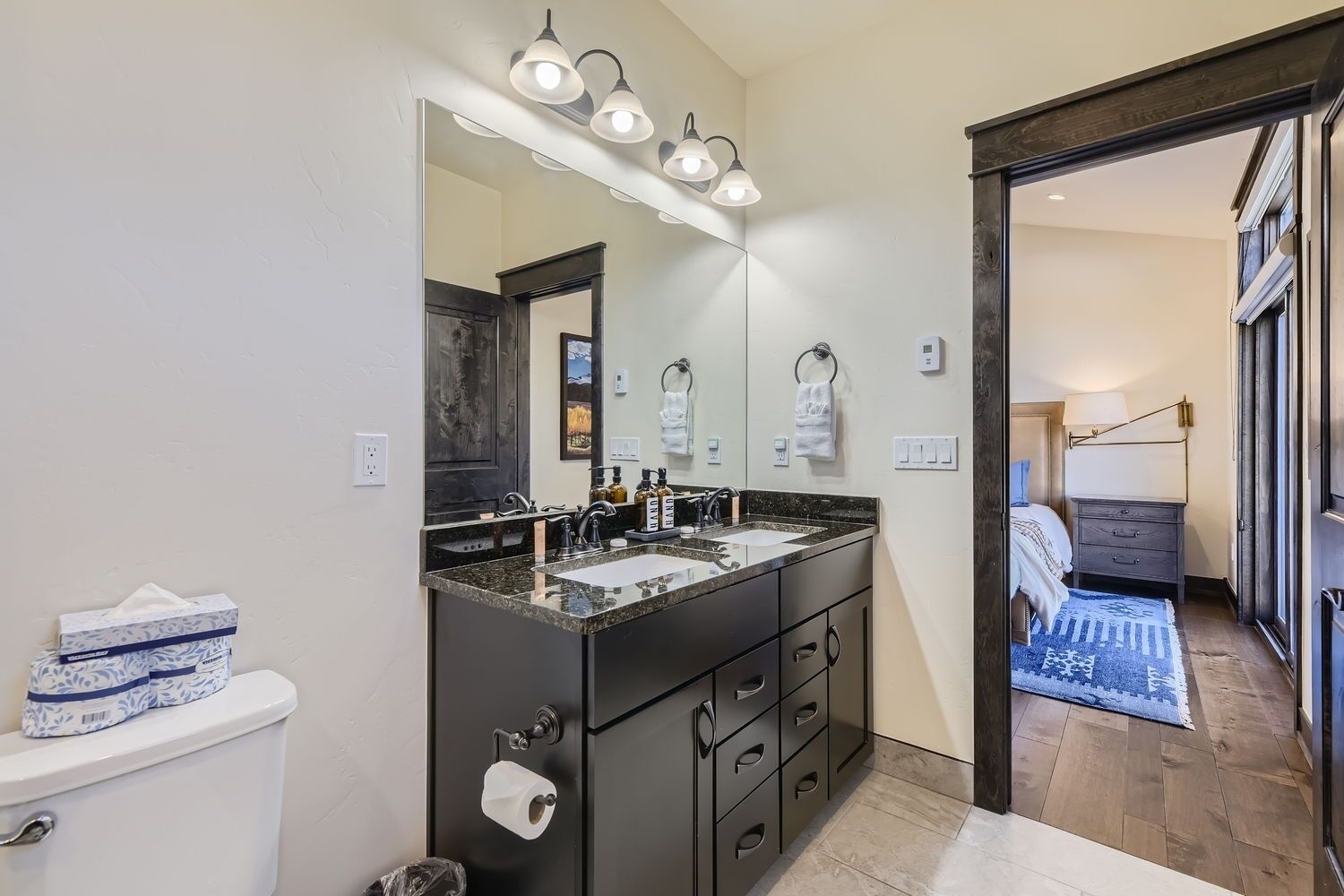 Un baño moderno con un mueble de tocador oscuro, dos lavamanos y un espejo grande. Se observan un dispensador de papel higiénico y un cubo de basura.