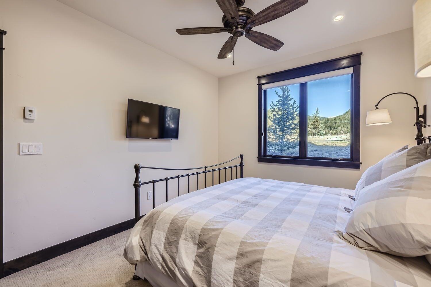Una camera da letto con un letto, una ventola a soffitto, una TV, una finestra vista alberi e una lampada.