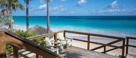 A wooden deck with beach chairs and a thatched roof structure overlooking a beach with clear turquoise water and palm trees.