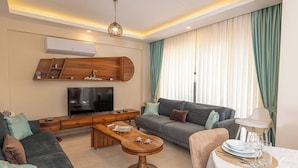 A modern living room with a flat-screen TV, wooden entertainment unit, grey sofa, and a dining area with a table set for two. - Close to Oludeniz 5 Bedroom Villa (Fethiye)