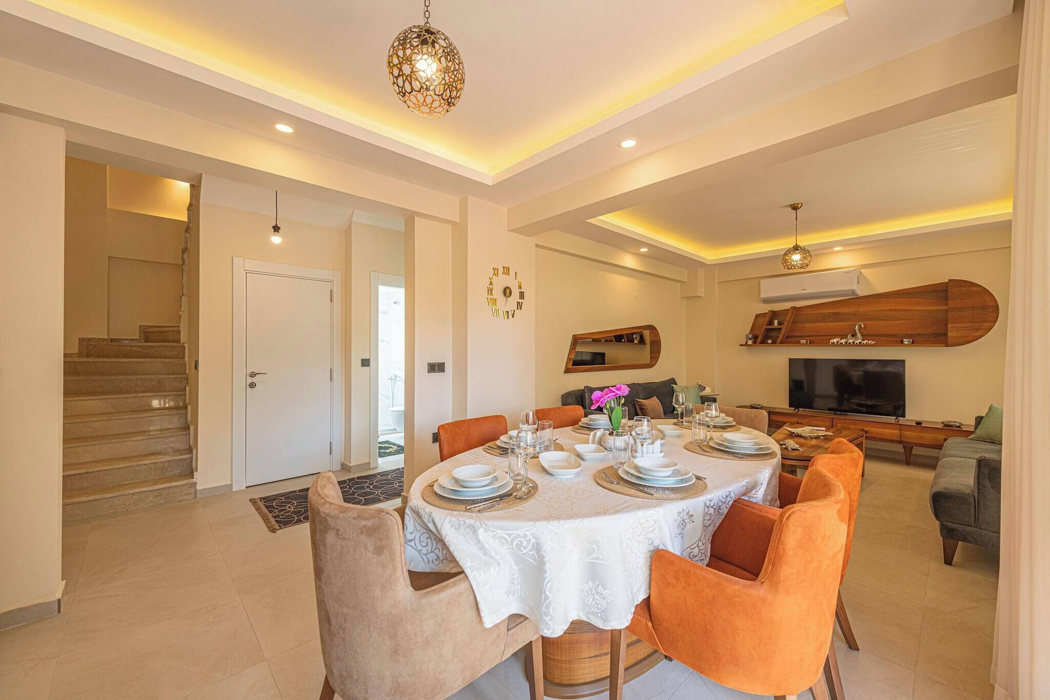 A dining area with a round table set for six, surrounded by orange chairs. There is a staircase leading to another level, a flat-screen TV mounted on the wall, and a wooden cabinet below the TV.