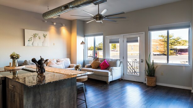 A spacious living area with a kitchen island, a ceiling fan, and large windows offering views of the outside.