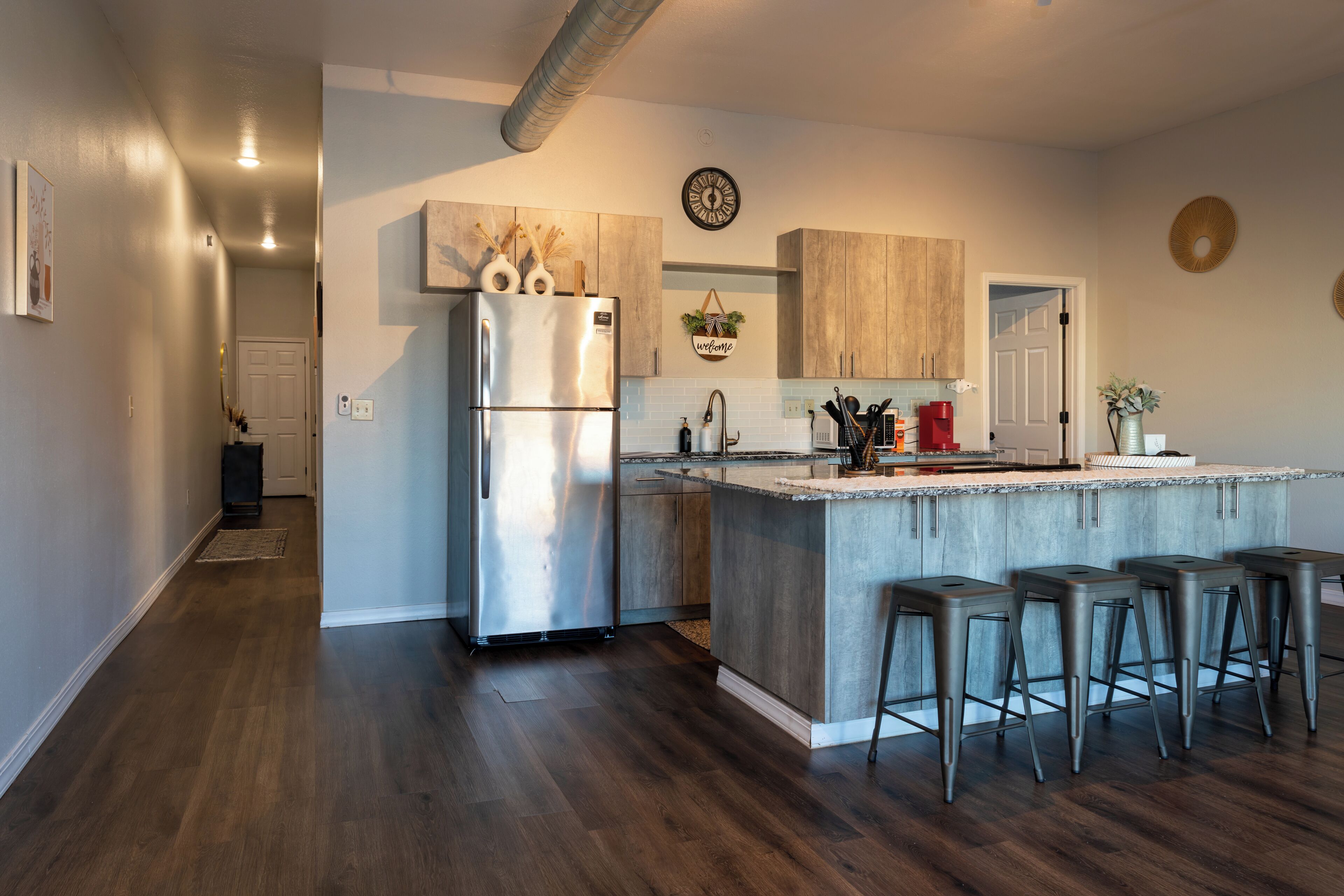 Una cucina moderna con frigorifero in acciaio inossidabile, pensili in legno, un angolo bar con sgabelli e un cartello che recita 'welcome'.