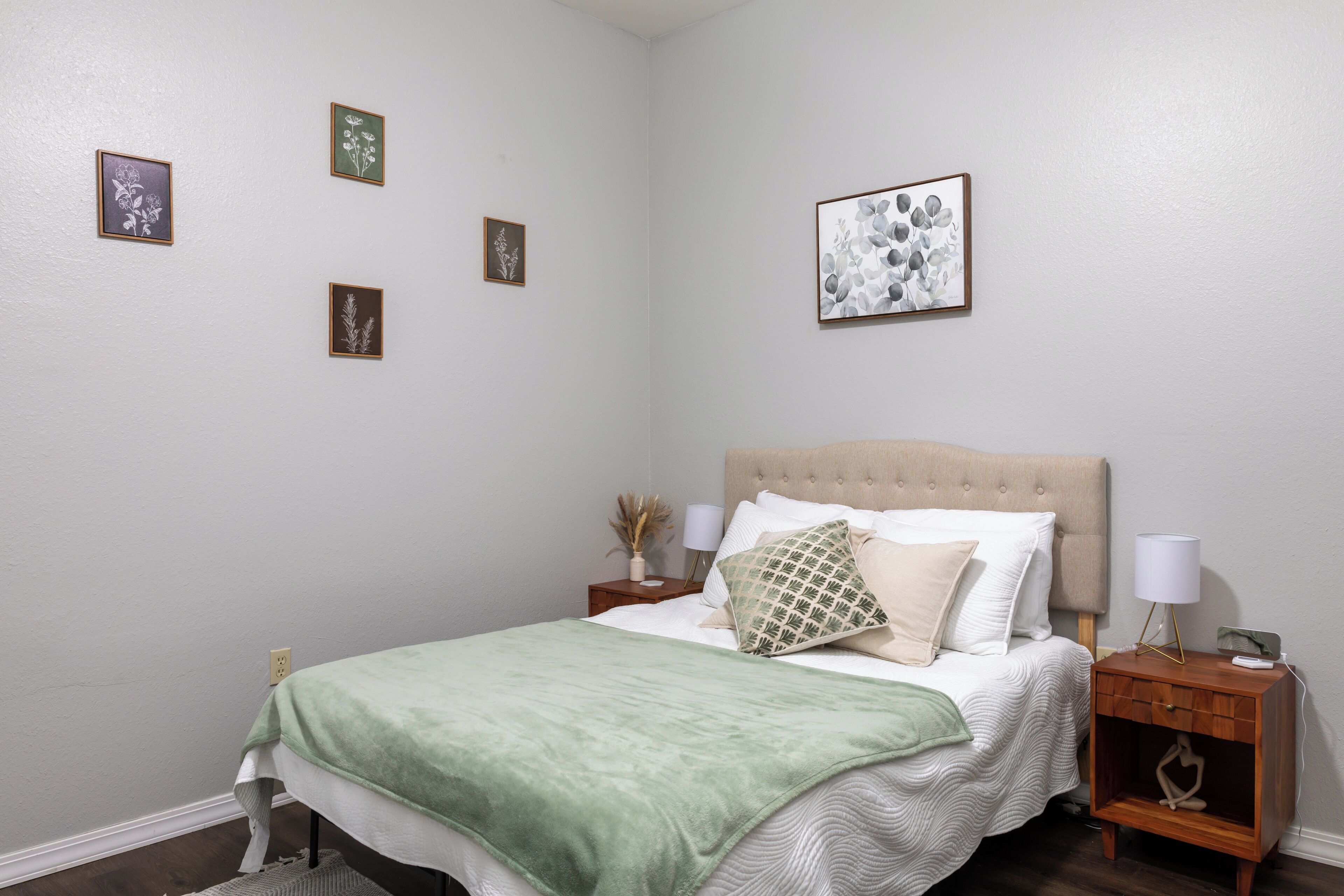 Un dormitorio con una cama, mesitas de noche, una lámpara y cuadros en la pared.