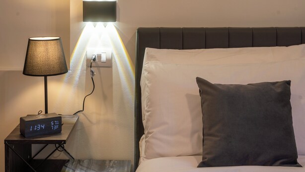 A neatly made bed with a pillow, a bedside lamp, and a digital clock on a small table beside the bed.