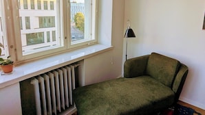 Una habitación con un ventanal grande, un radiador, un sofá verde, una lámpara de pie negra y una alfombra estampada.