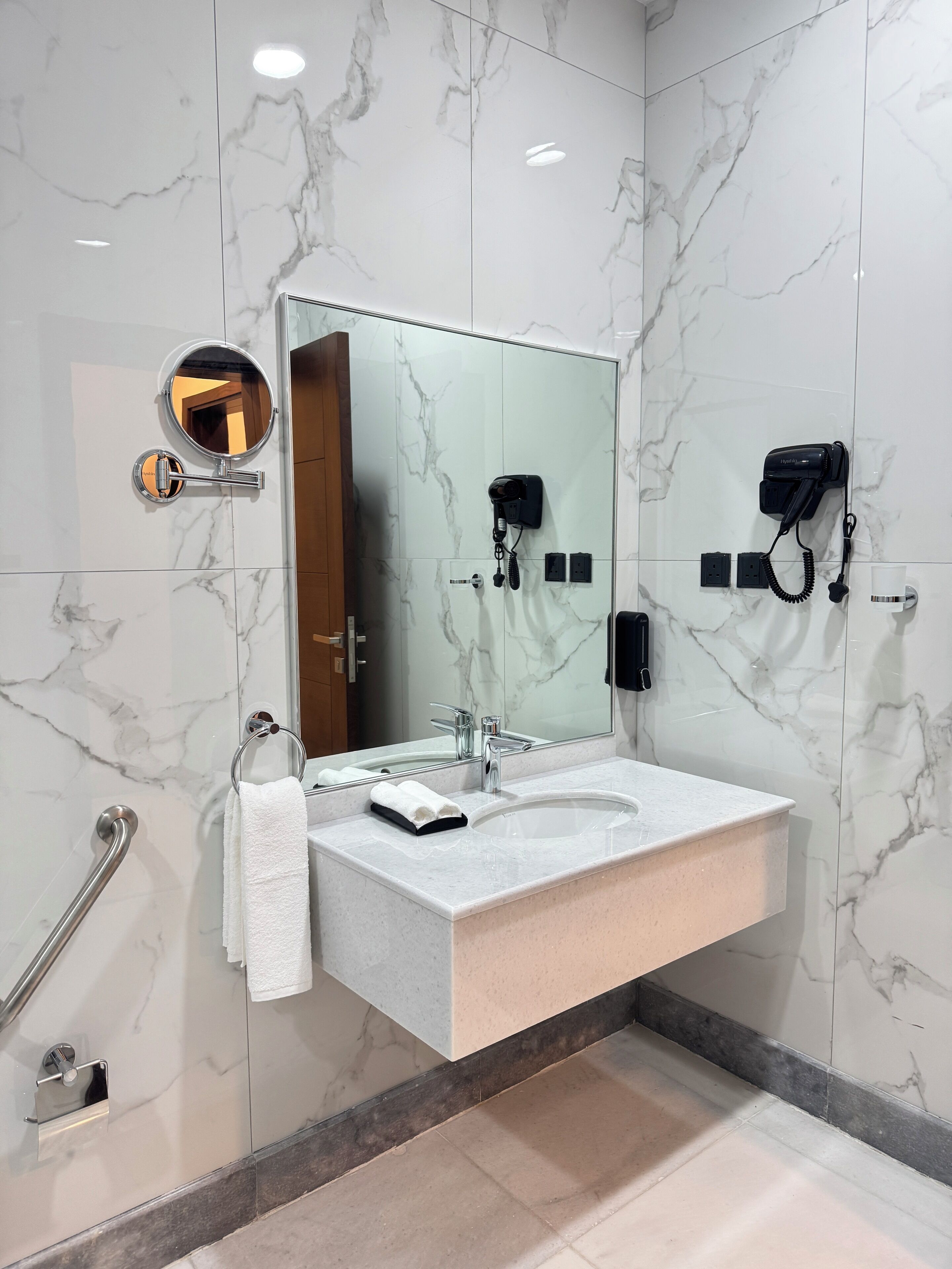 Une salle de bain moderne avec un mur en marbre, un grand miroir, un lavabo équipé d’un combiné téléphonique et un sèche-cheveux fixé au mur.
