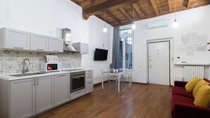 A compact kitchen with a microwave, oven, and sink, a dining area with a table and chairs, and a red sofa in a living area with a wooden ceiling.