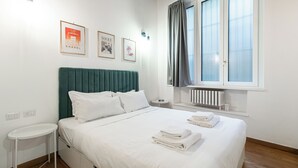 A neatly made bed with white linens and pillows, a bedside table, a radiator below a window with curtains, and framed pictures on the wall.