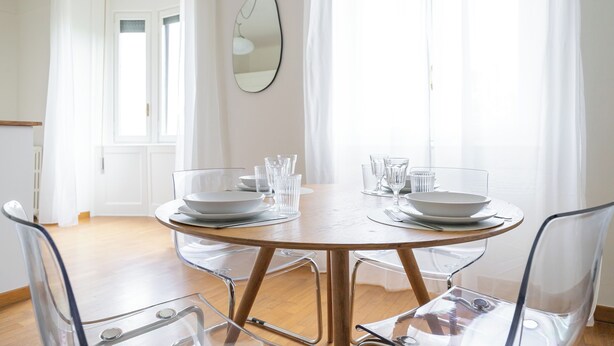 Une table de salle à manger ronde, entourée de chaises transparentes, avec de l’argenterie blanche et des verres, le tout disposé dans une pièce lumineuse dotée d’un plancher en bois et de murs blancs.