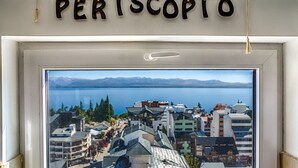 Ein Fenster mit Blick auf eine Küstenstadt, ein Schild darüber mit der Aufschrift „PERISCOPIO“ und ein weißer Rahmen mit einem kleinen weißen Gegenstand in der Mitte.