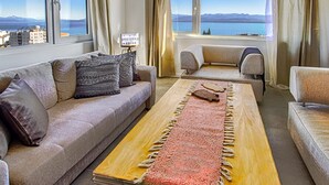 Ein Wohnzimmer mit einer Couch, Sesseln und einem Couchtisch. Zwei Fenster mit Vorhängen, und ein Schild ist auf dem Fensterbrett sichtbar.
