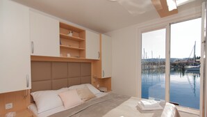 Une chambre à coucher comprenant un lit, des tables de chevet, une tête de lit et une vue sur une marina, grâce à de grandes fenêtres.