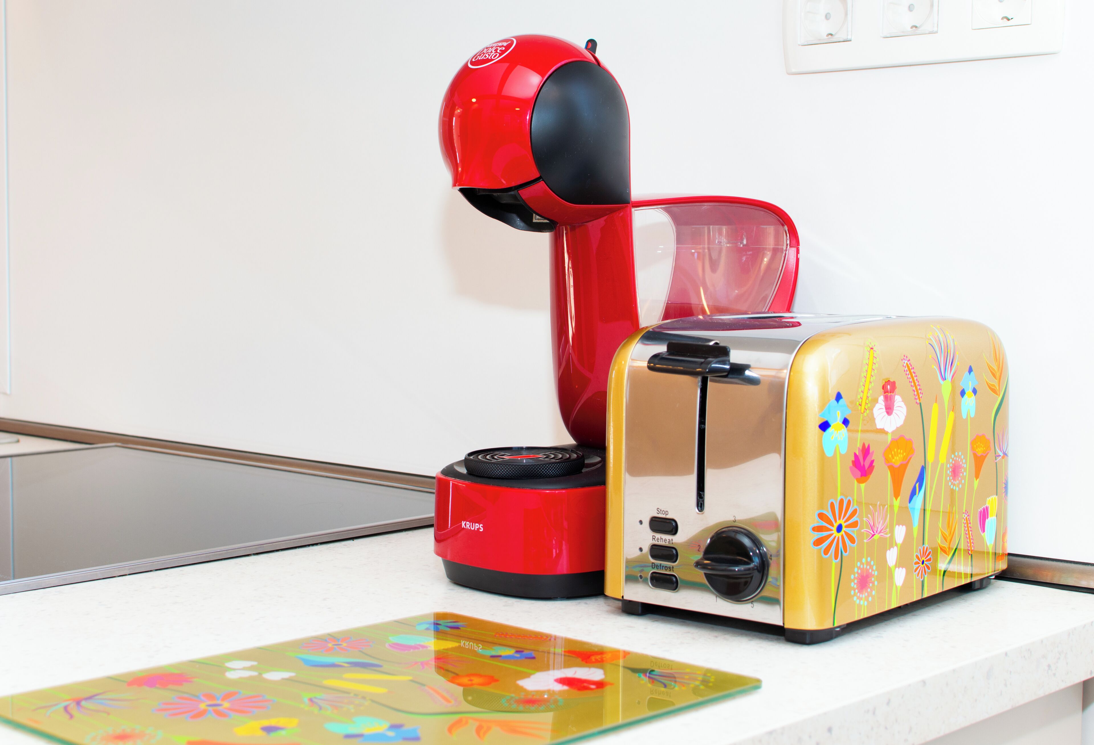 Ein roter und schwarzer Wasserkocher, ein Toaster mit Blumenmuster und eine farbenfrohe Matte auf einer Küchenarbeitsfläche.