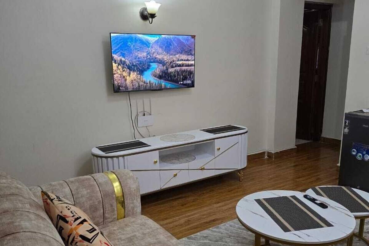 Ein Wohnzimmer mit flachbildfernseher an der Wand, einer weißen Unterhaltungseinheit, einem grauen Sofa mit goldenen Akzenten und einem kleinen runden Couchtisch.