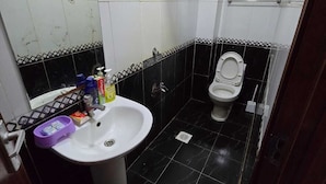 A small bathroom with a white sink, a toilet, and a mirror. There are toiletries on the sink and a shelf with cleaning supplies above the toilet. - Lux Suites Astrol Apartments Garden City (Nairobi)