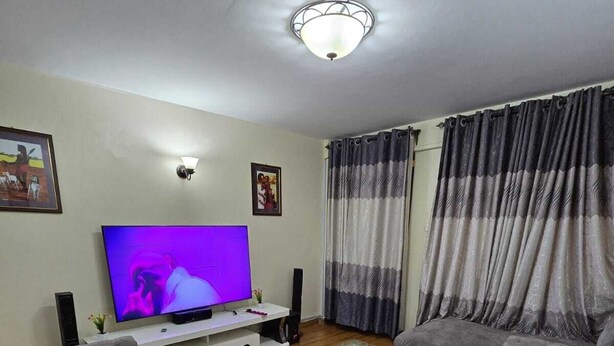 A living room with grey sofas, a glass coffee table, a flat-screen TV, and a ceiling light fixture. - Lux Suites Astrol Apartments Garden City (Nairobi)
