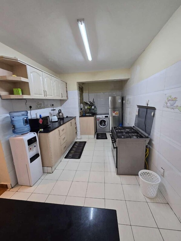 A compact kitchen with white cabinets, a water dispenser, a stove, and a washing machine. - Lux Suites Astrol Apartments Garden City (Nairobi)