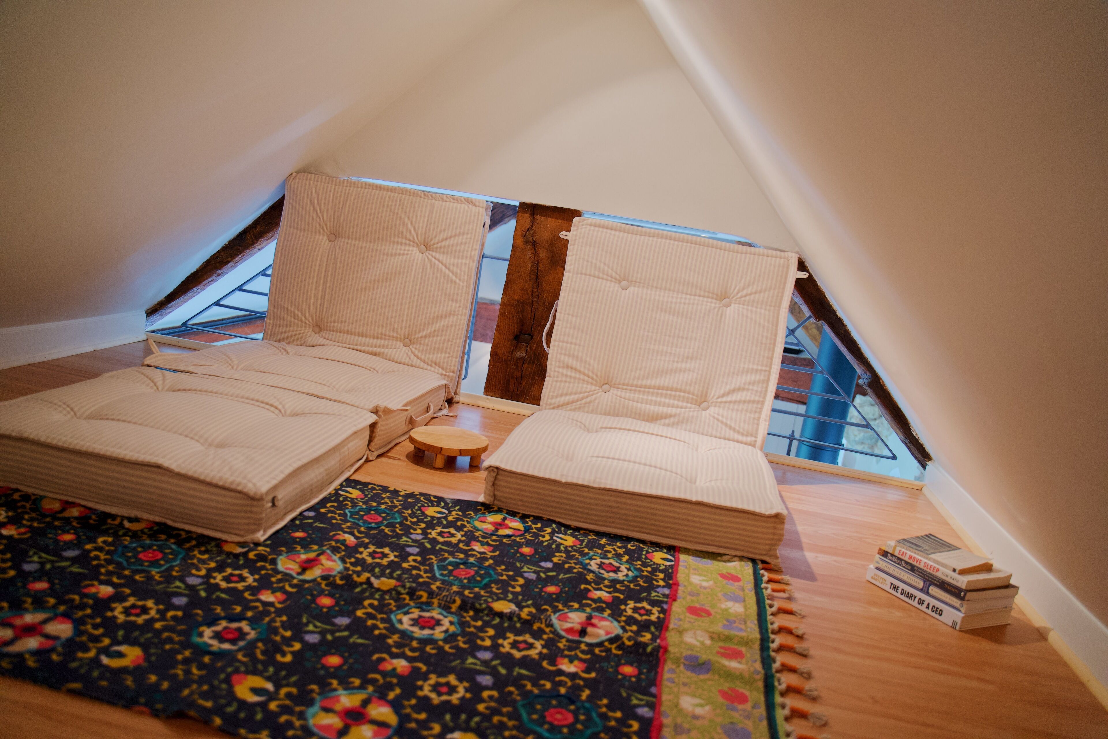 Une chambre mansardée chaleureuse avec un plafond en pente, deux coins salon confortables, un petit tabouret en bois et un tapis coloré.