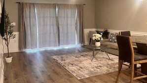 Ein modernes Wohnzimmer mit Essbereich, Holzboden, einem großen Fenster mit Vorhängen, einem Topf mit Pflanze und einem Glastisch.