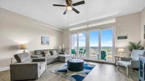 A spacious living room with a grey sectional sofa, a round ottoman, and a ceiling fan.