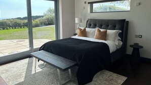 A modern bedroom with a large bed, a bench, and a view of a green landscape through large windows. - Spacious 6-bedroom house with WiFi, AC in enjoyable Dunmore East (Dunmore East)