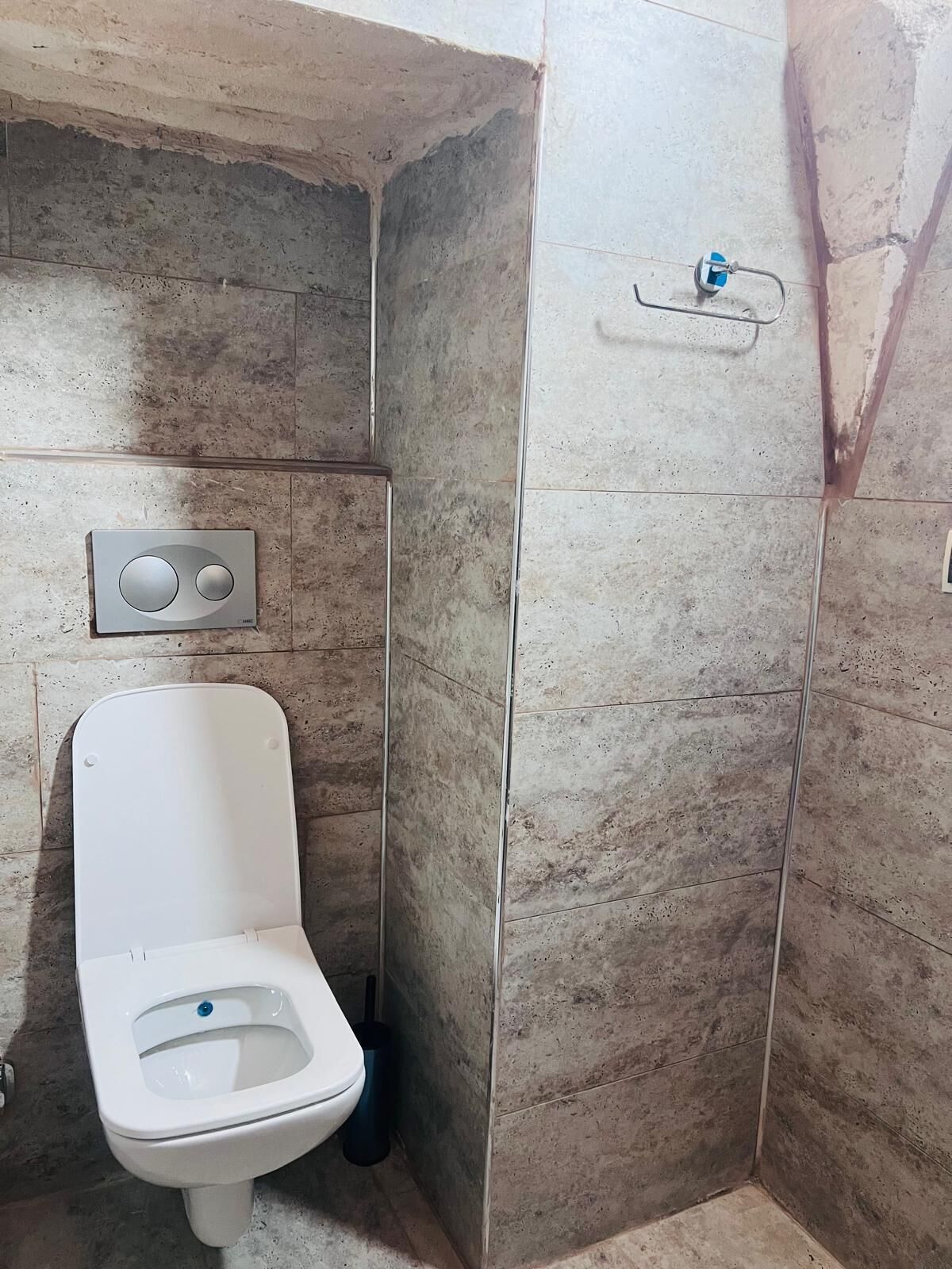 Un baño pequeño con paredes de azulejos, un inodoro blanco, un accionador de descarga en la pared y una zona de ducha.